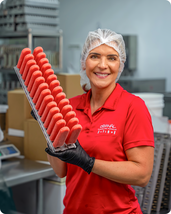 A smiling female food production worker wearing a red polo shirt, translucent hairnet, and black gloves holds a large metal tray filled with rows of red frozen popsicles inside a commercial production facility. Cardboard boxes, stainless steel equipment, and industrial machinery appear softly blurred in the background. A partially cropped decorative text graphic is visible in the upper-right corner.