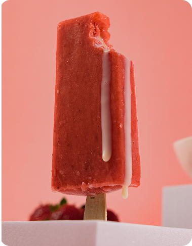 A close-up product image shows a red strawberry ice cream bar standing upright against a soft pink background. A bite is missing from the top corner, revealing the interior, while a creamy white filling drips vertically down the front and side. The wooden stick is visible at the bottom, with blurred strawberries and white geometric props in the background.